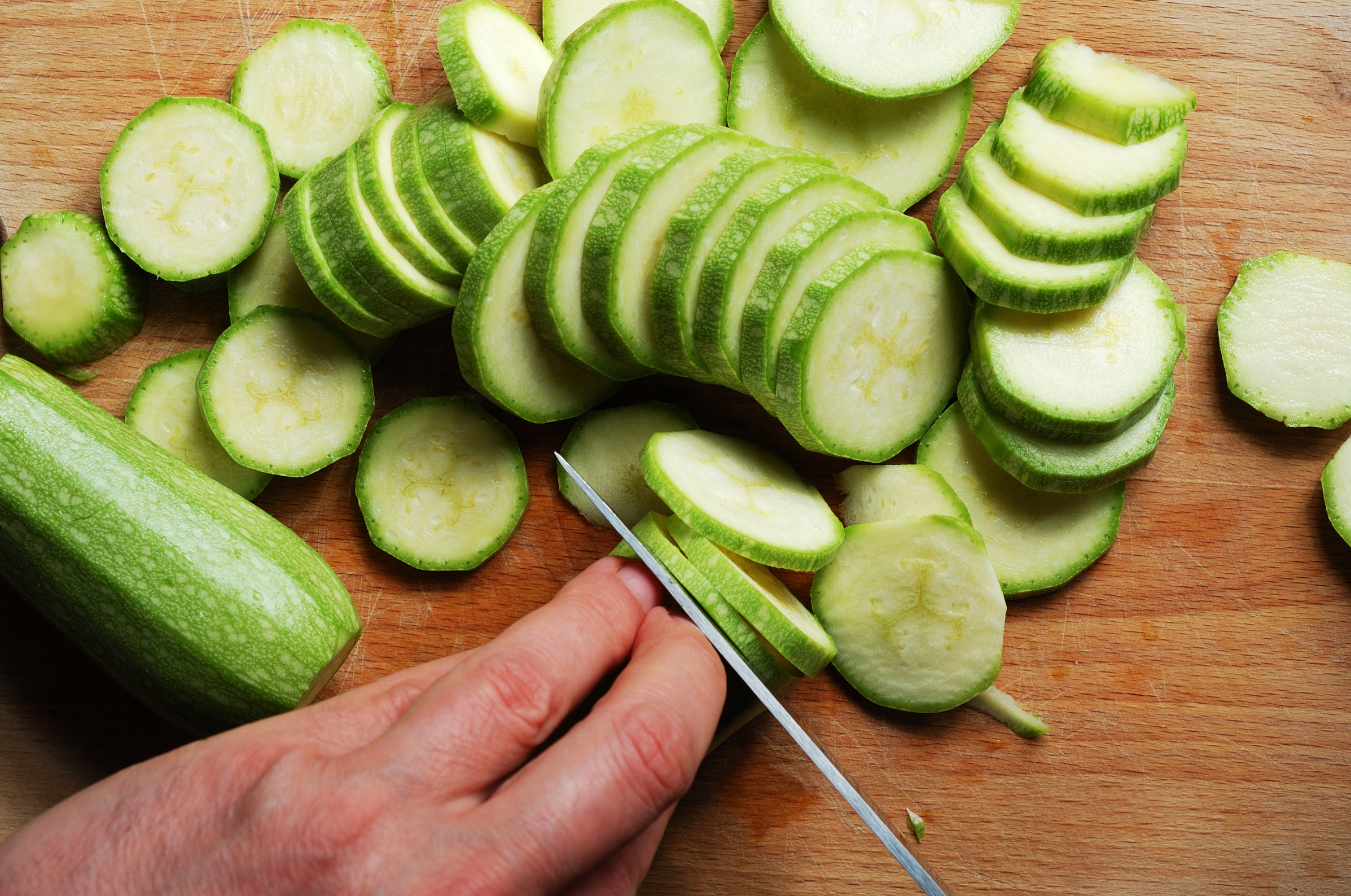 Hand slicing zucchini into thin rounds on a wooden cutting board with a Japanese knife.