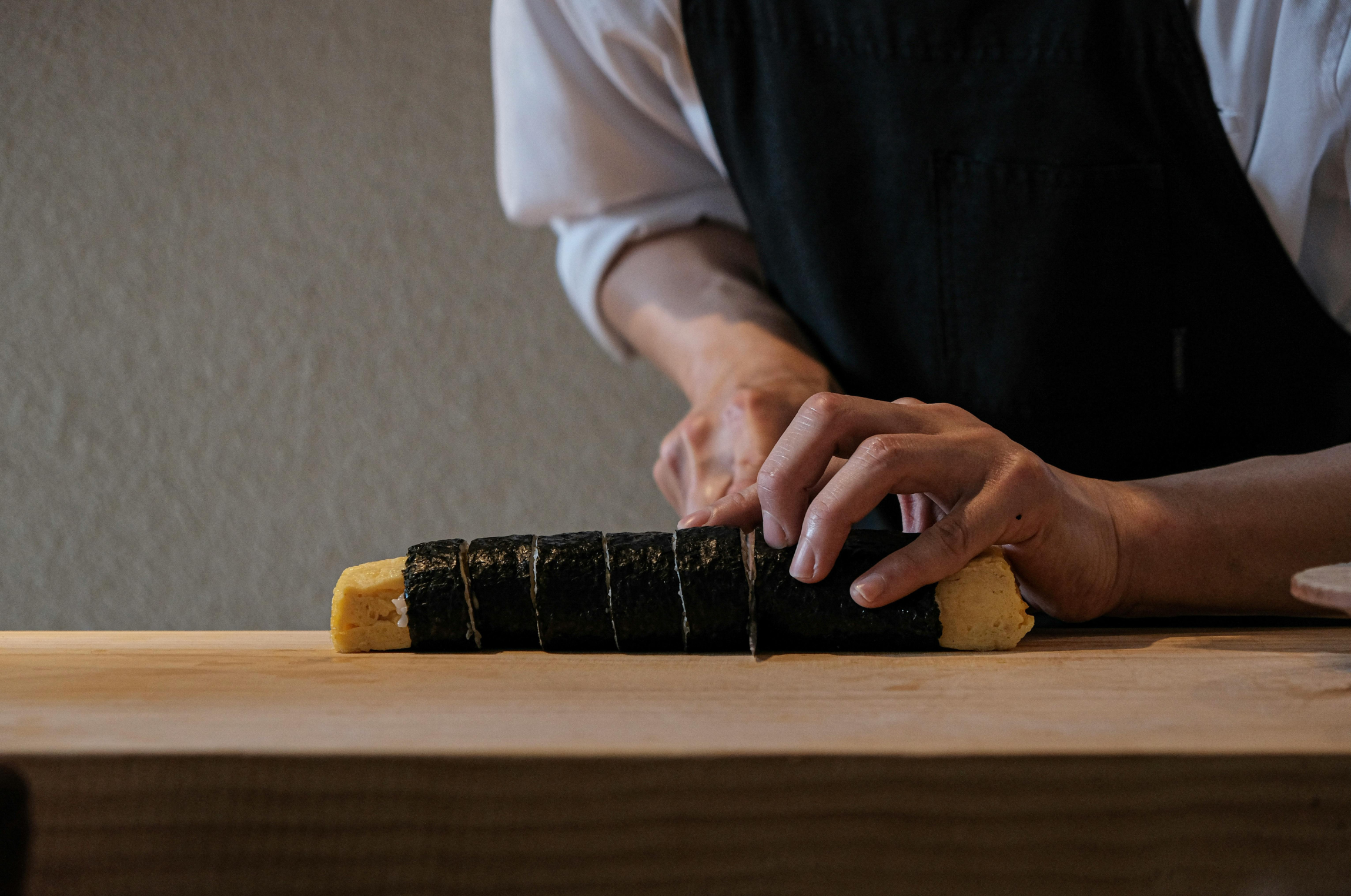 Image of Pro Chefly chef slicing a tamagoyaki sushi roll wrapped in nori on a wooden cutting board.