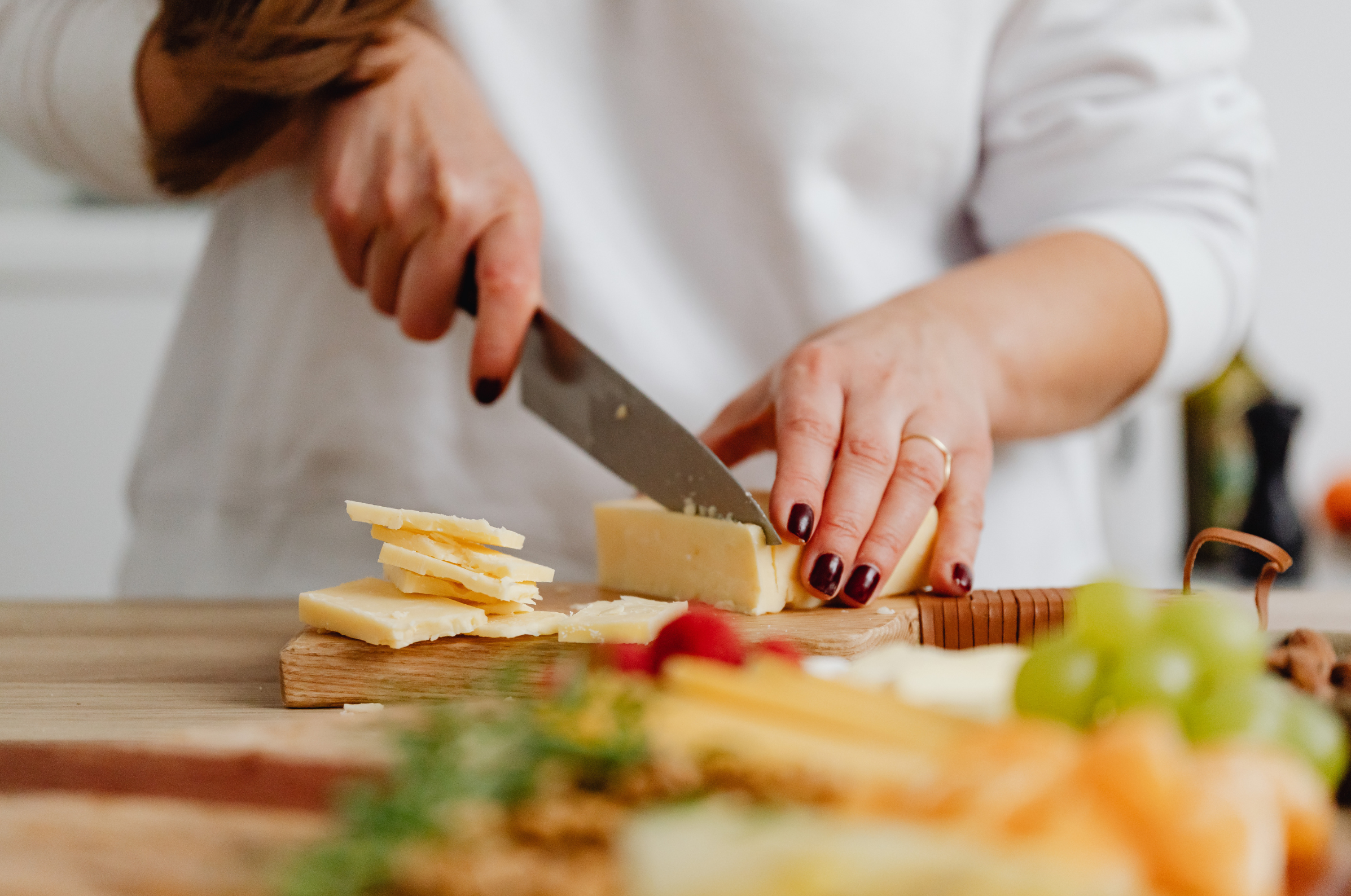 Image of Pro Chefly Japanese Damascus chef knife slicing aged cheese on a wooden board with precision, surrounded by fresh grapes, herbs, and charcuterie ingredients in a clean kitchen setting