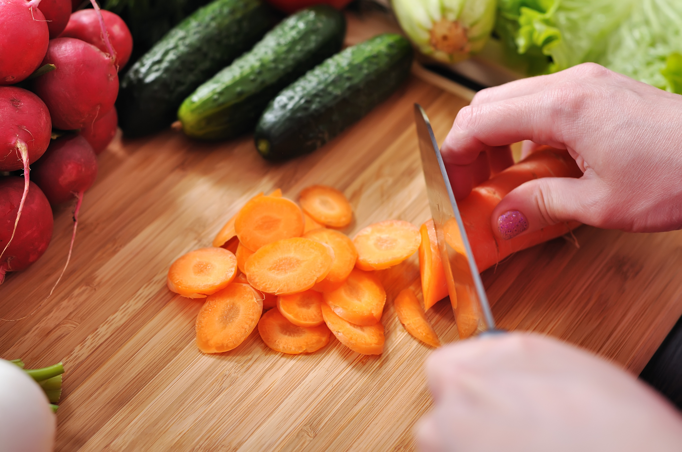 Image of Pro Chefly Japanese Damascus knife slicing fresh carrots on a wooden cutting board, surrounded by radishes, cucumbers, and leafy greens during vegetable prep.