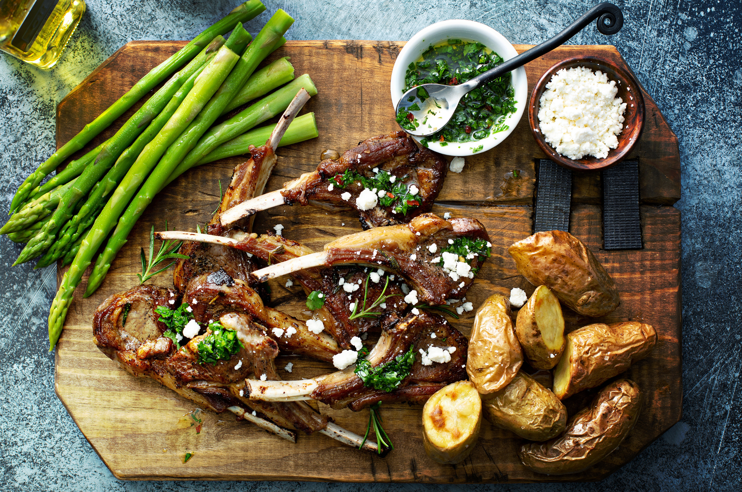 Image of Pro Chefly mint-crusted lamb chops on a wooden board with roasted potatoes, asparagus, chimichurri sauce, and crumbled cheese, styled in an overhead gourmet presentation with vibrant, natural lighting