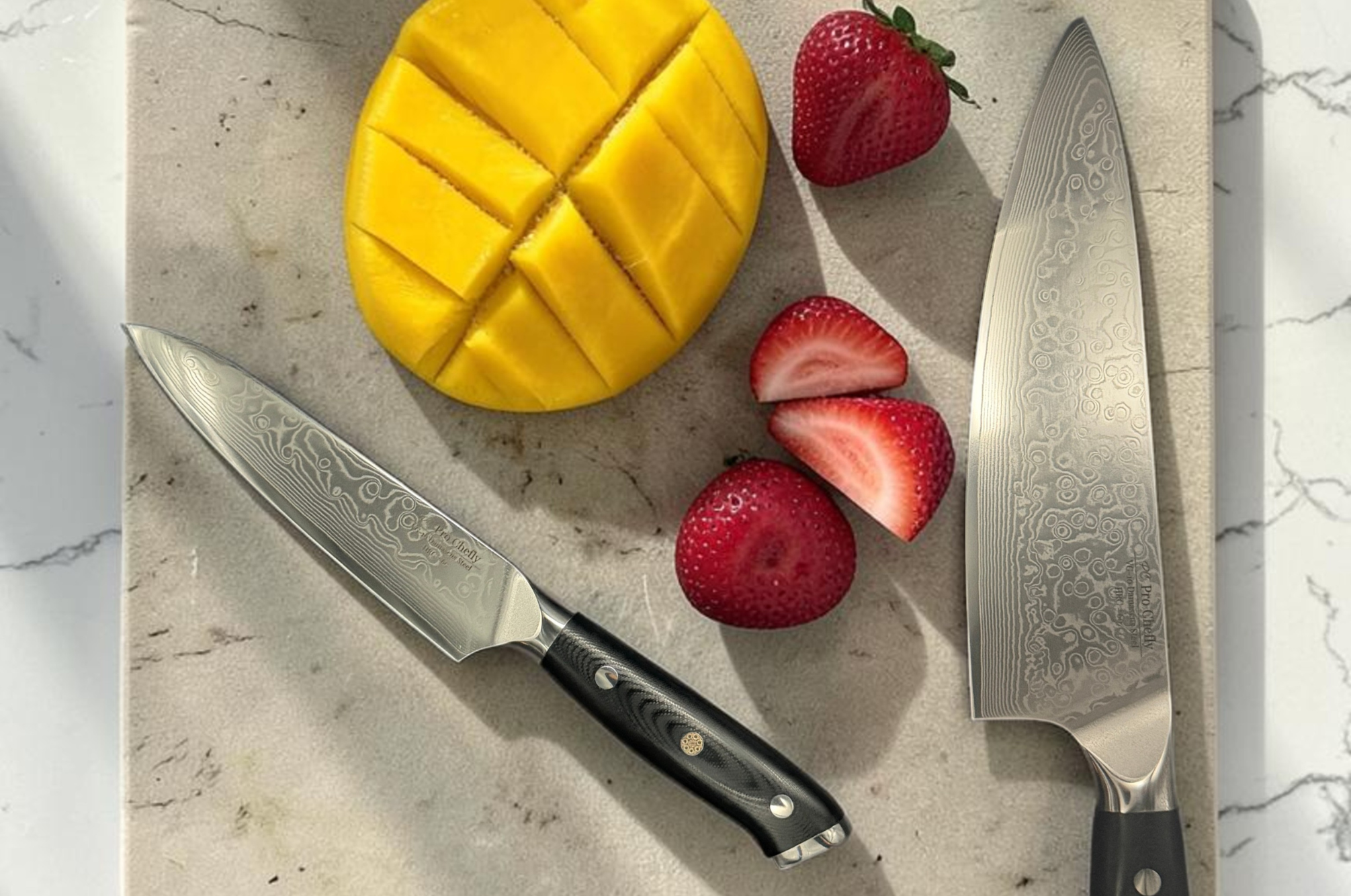 Image of Pro Chefly Japanese Damascus chef knife and paring knife beside fresh mango and sliced strawberries on a marble countertop, premium kitchen prep photography highlighting precision cutlery.