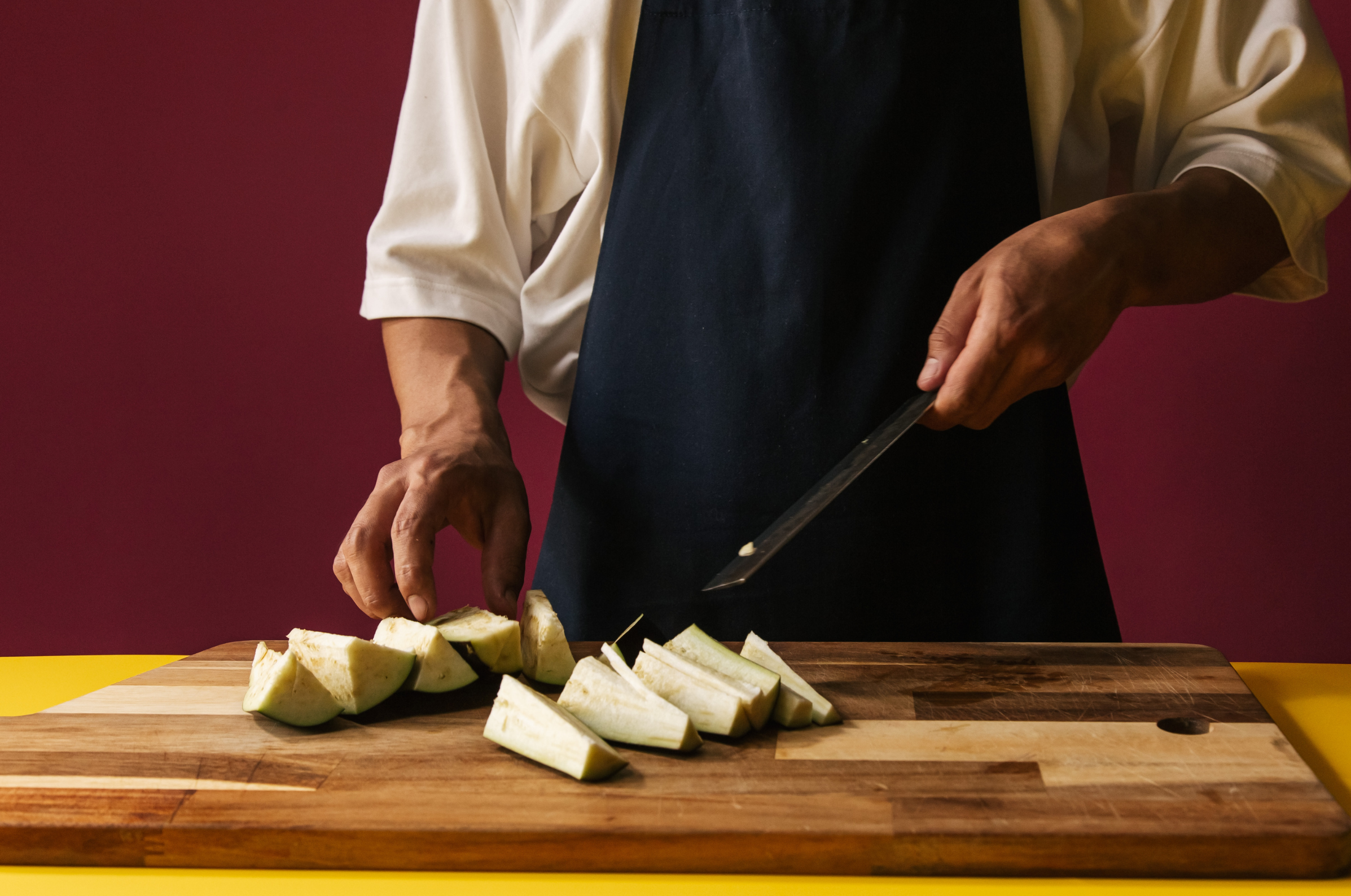 Image of Pro Chefly chef slicing eggplant wedges on a wooden cutting board with a kitchen knife during meal preparation.