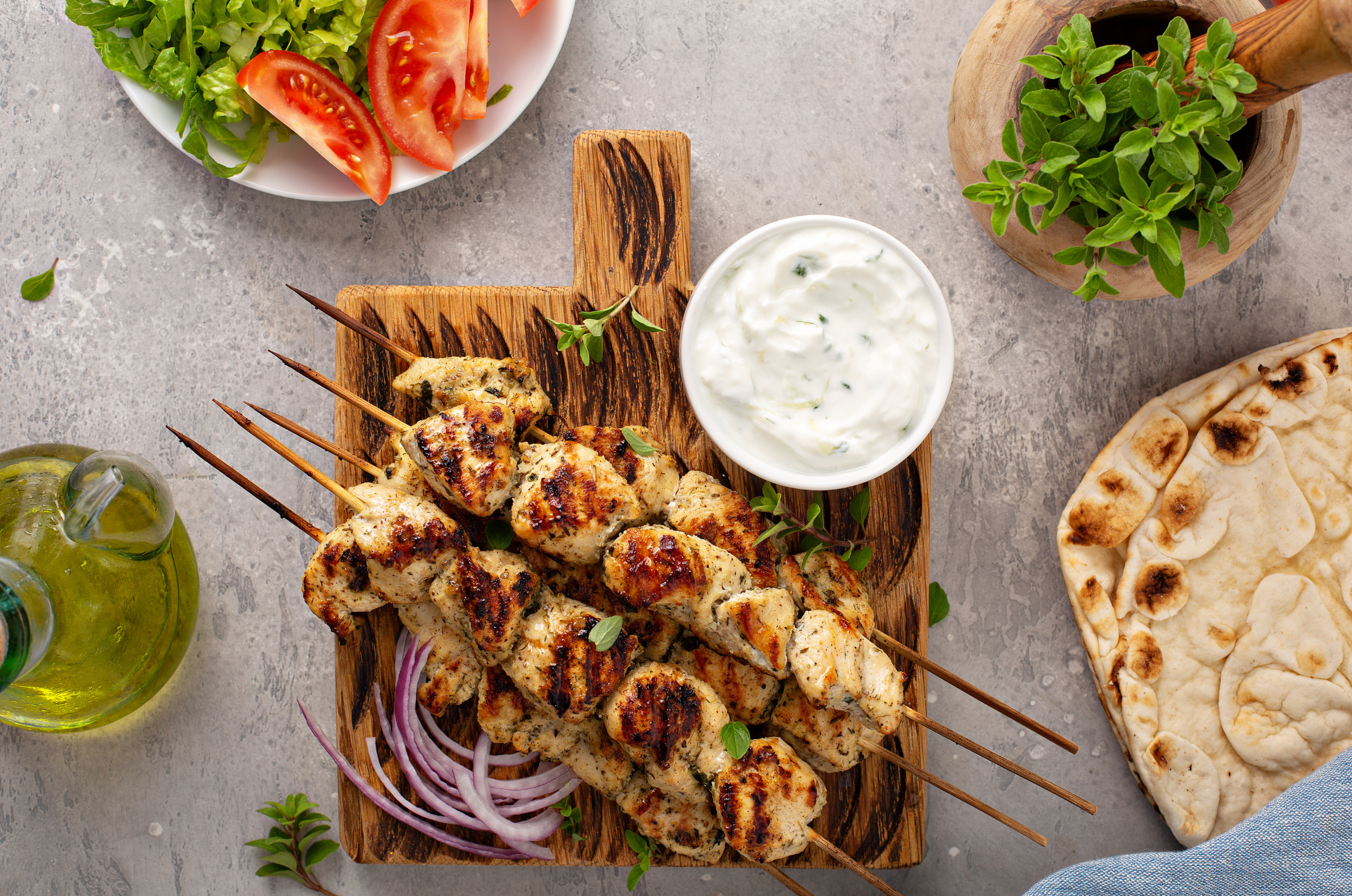 Image of Pro Chefly chicken souvlaki served with creamy herb yogurt sauce, warm naan bread, fresh tomatoes, lettuce, red onion, and oregano on a rustic wooden board.