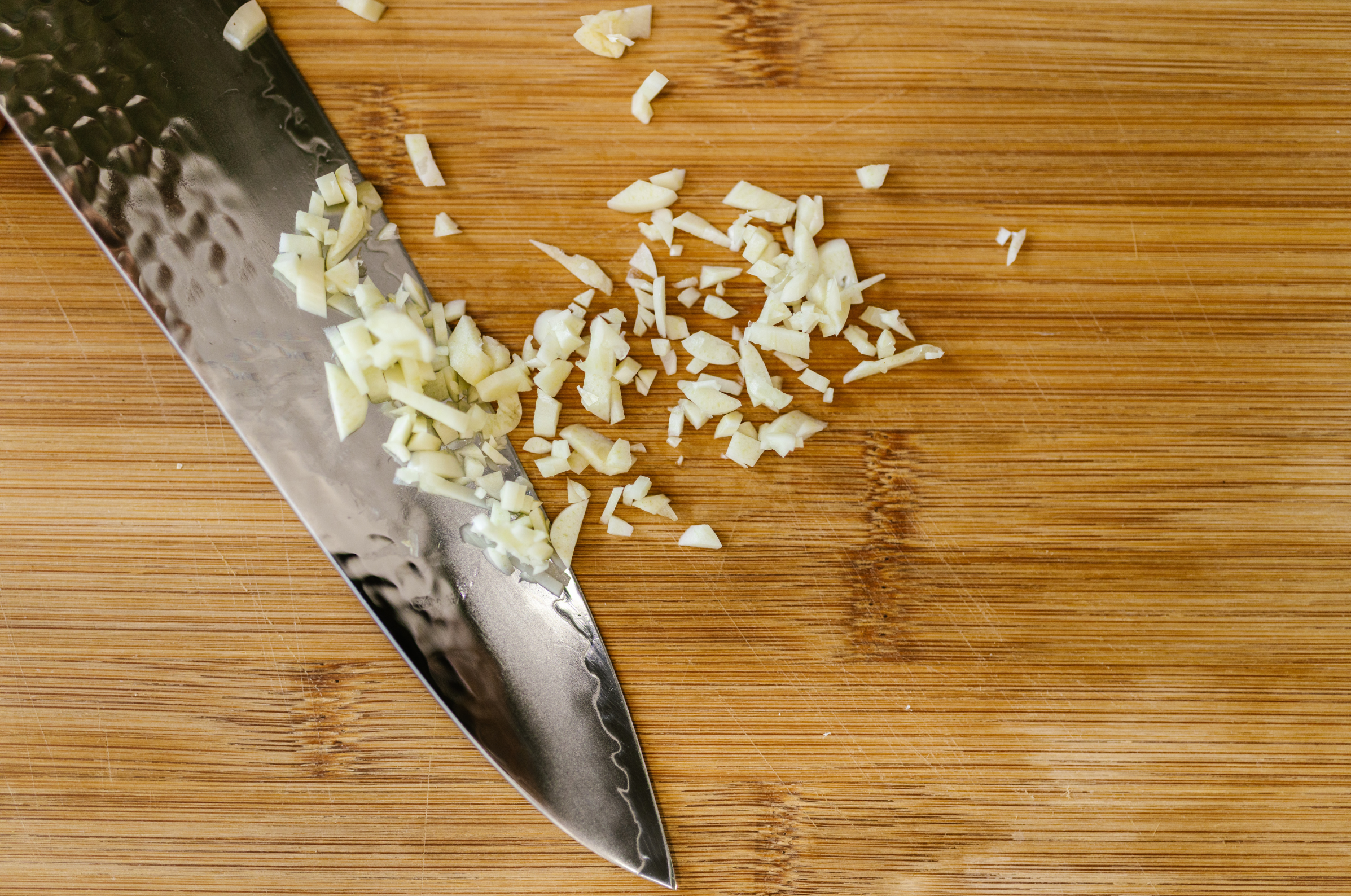 Image of Pro Chefly Damascus chef knife finely mincing garlic on a wooden cutting board, showcasing sharp Japanese knives precision and controlled prep technique.