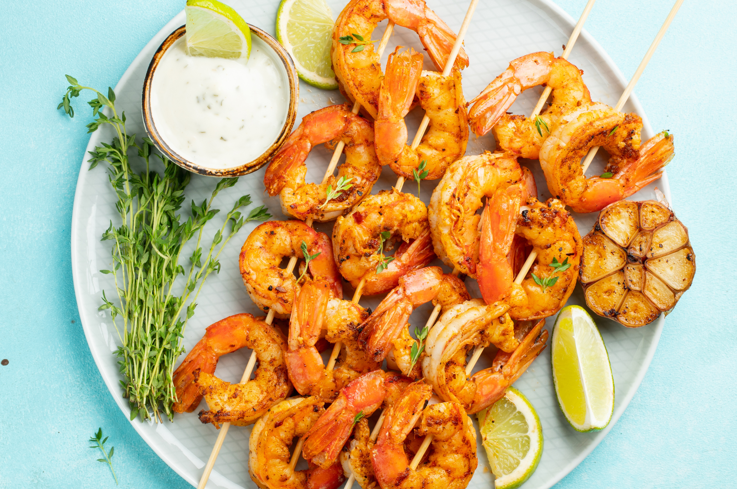 Image of Pro Chefly grilled shrimp skewers with lime wedges, roasted garlic, fresh thyme, and creamy dipping sauce on a serving platter.