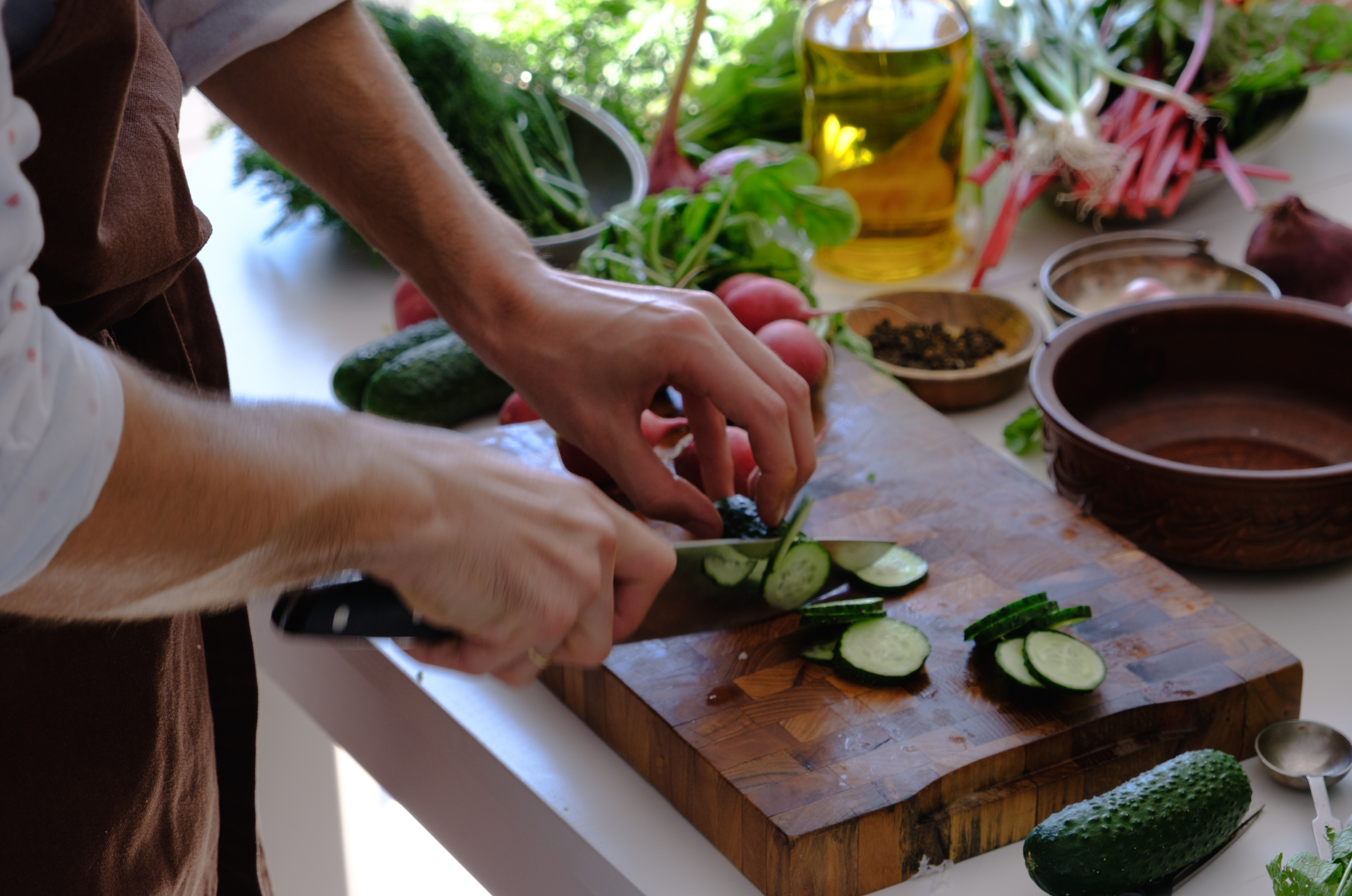 Image of Pro Chefly chef slicing fresh cucumbers with a sharp Japanese knife on a wooden cutting board, surrounded by garden vegetables and olive oil in a bright kitchen.