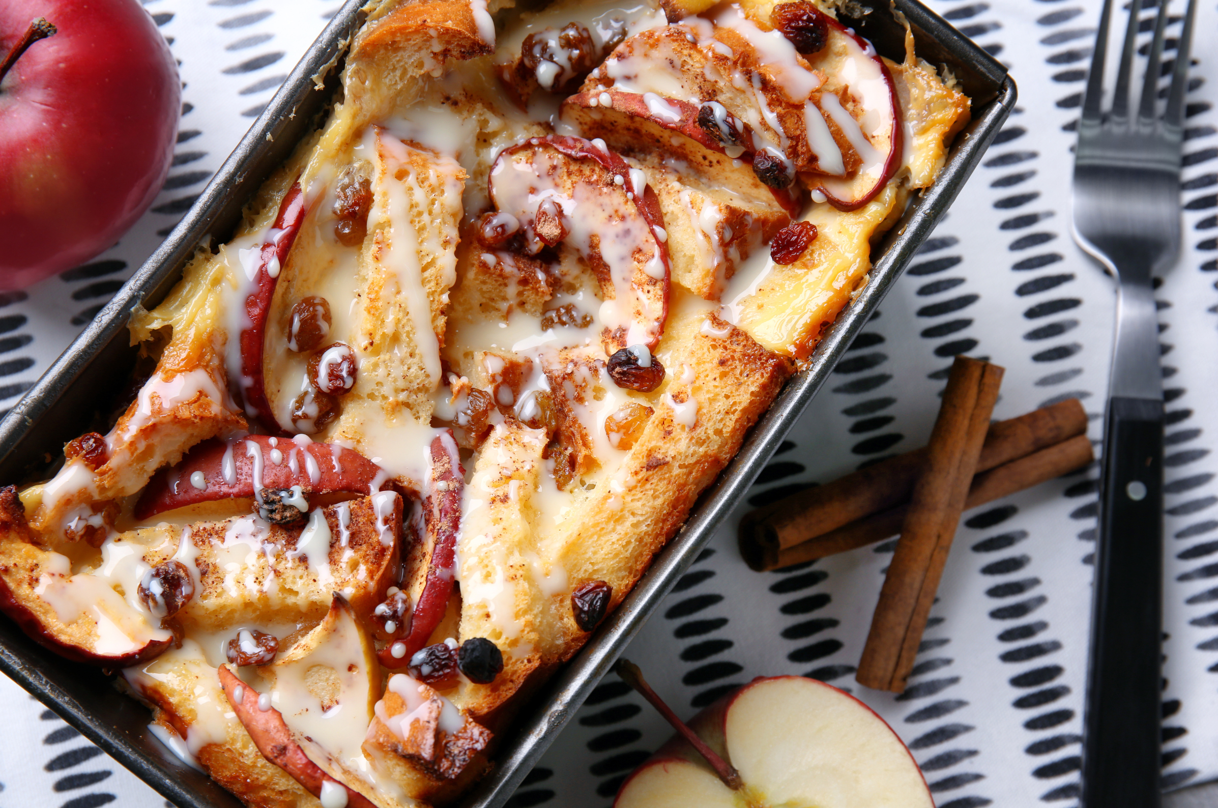 **Alt text:** Apple bread pudding with raisins and icing in a baking dish beside fresh apples and cinnamon sticks.