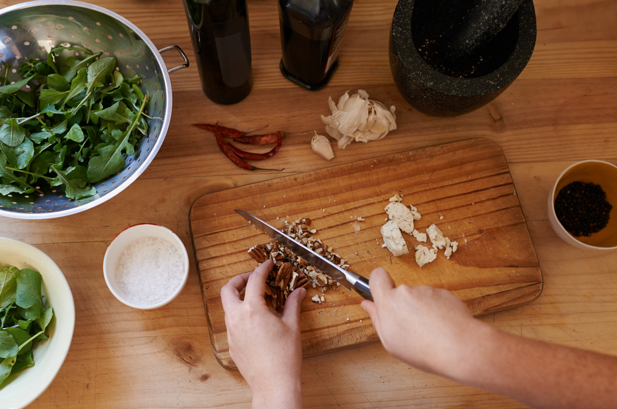 Image of Hands chopping pecans on a wooden board with greens, spices, and cheese nearby.