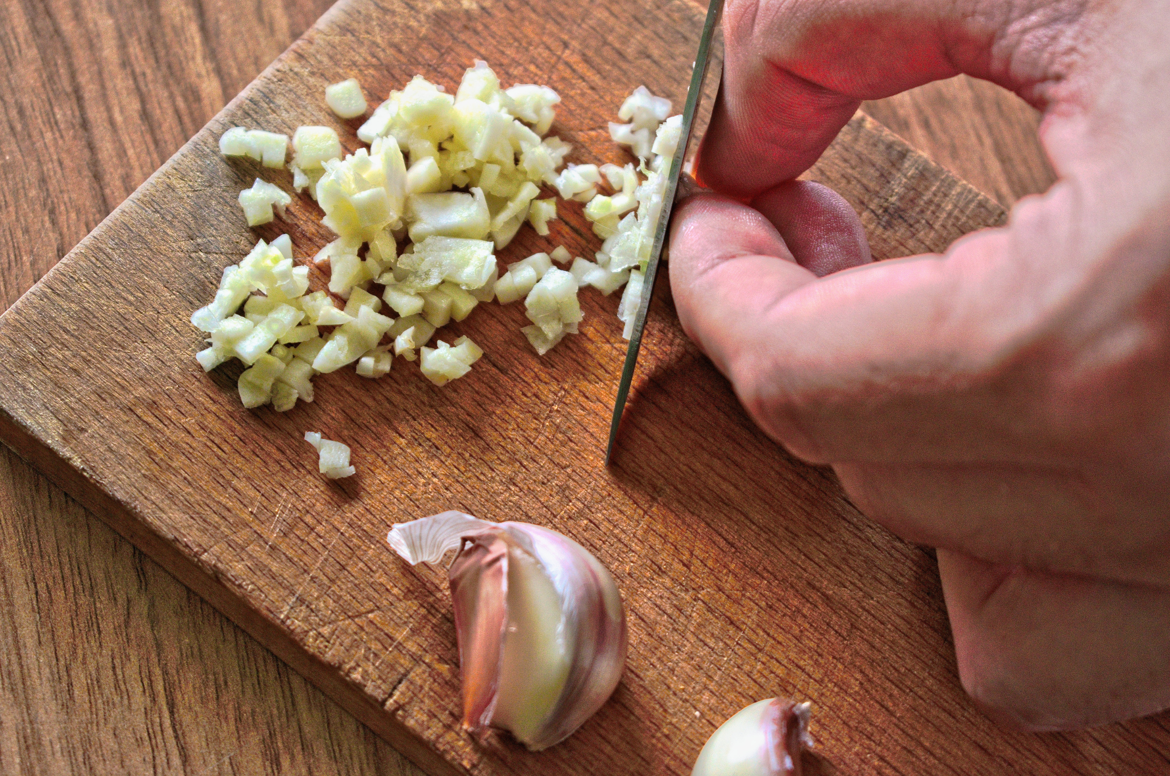 Image of pro chefly damascus knife finely mincing fresh garlic on wooden board, emphasizing precision cutting and culinary craftsmanship