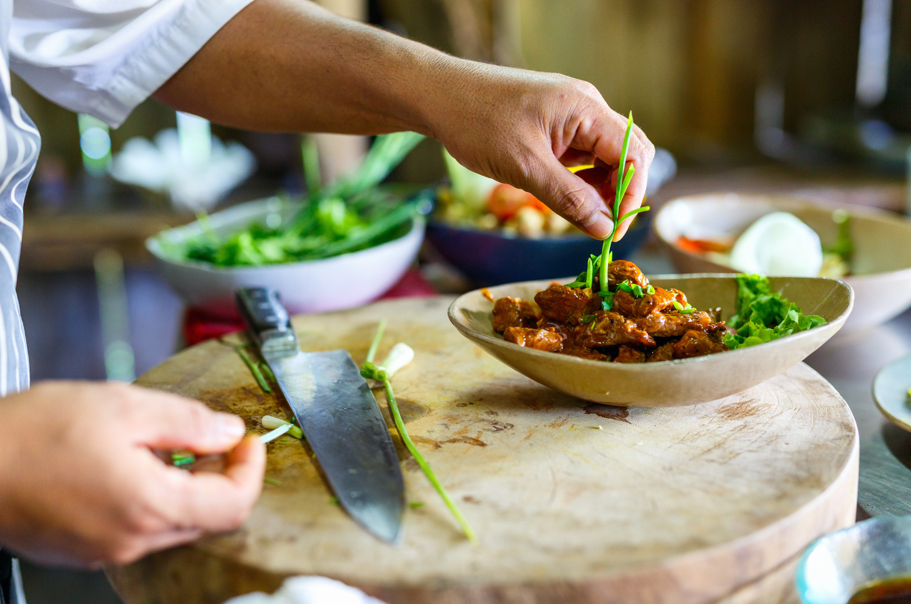 Chef garnishing a bowl of seasoned meat with fresh herbs on a wooden board, Pro Chefly knife nearby, showcasing precision and plating artistry.