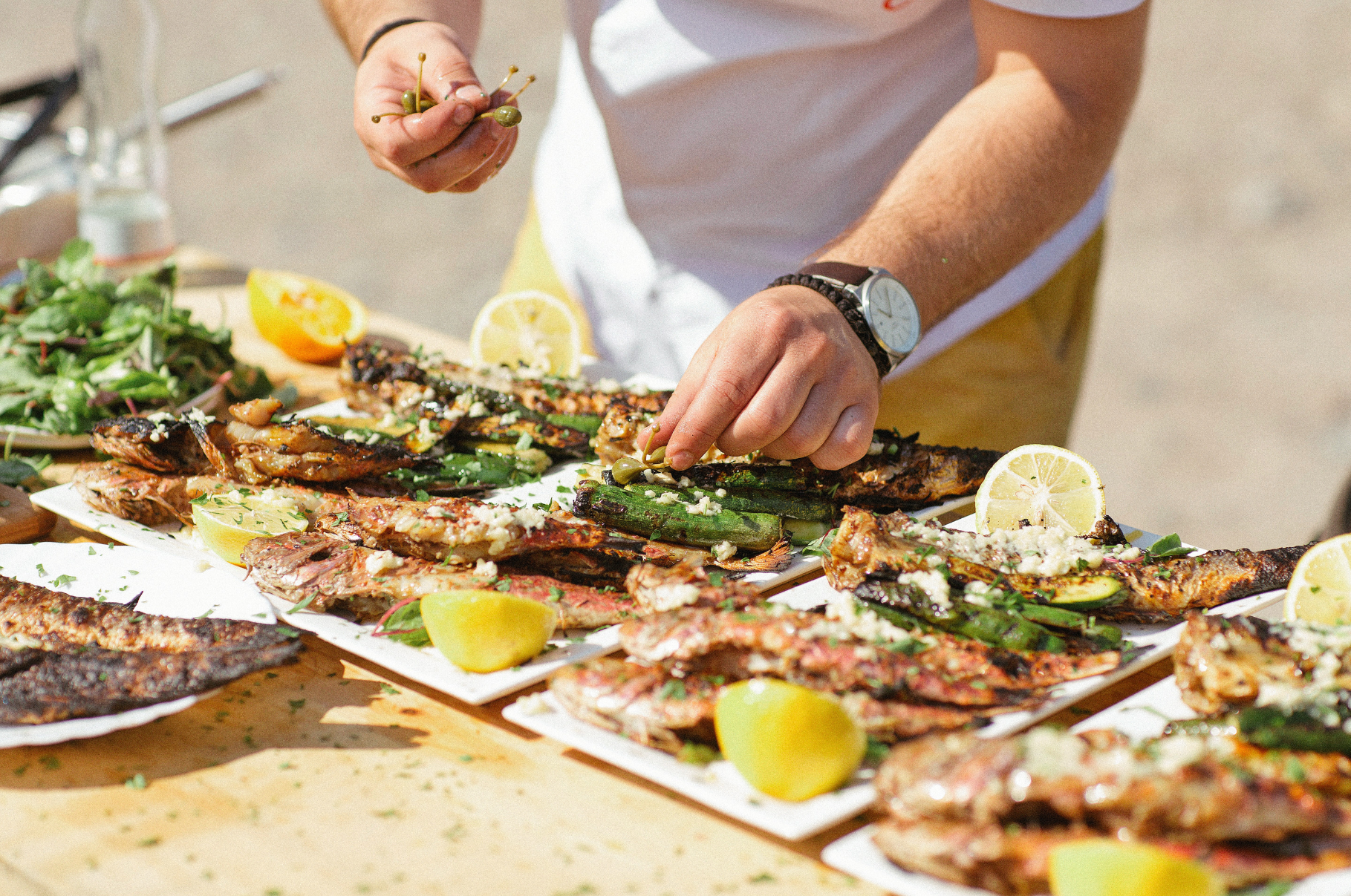 Chef garnishing grilled fish with fresh toppings and lemon slices outdoors, creating a vibrant seafood spread styled for Pro Chefly’s culinary storytelling.