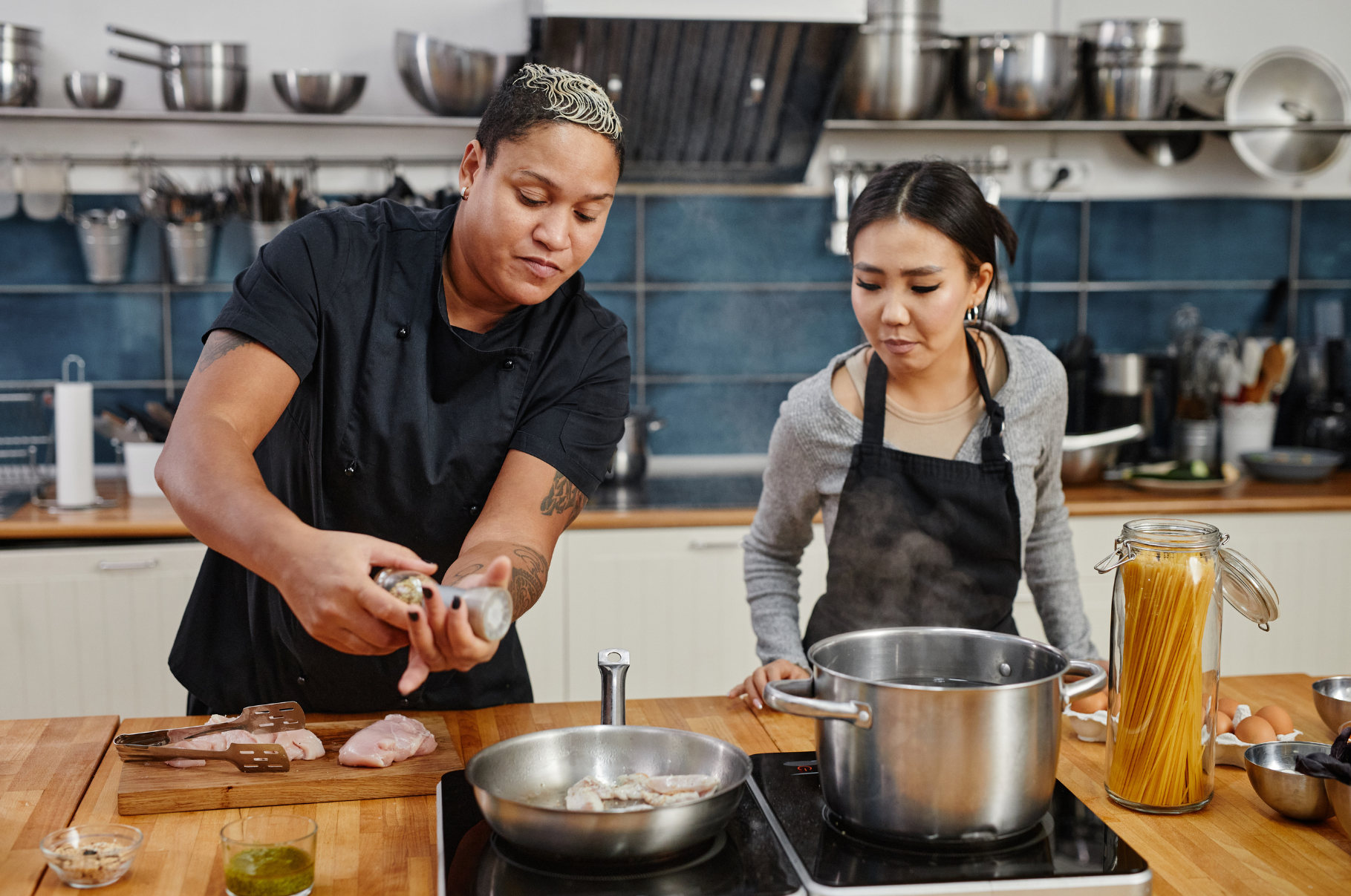 Two chefs cooking together in a modern kitchen, one seasoning chicken in a pan while the other watches pasta boil, showcasing teamwork and Pro Chefly culinary style.