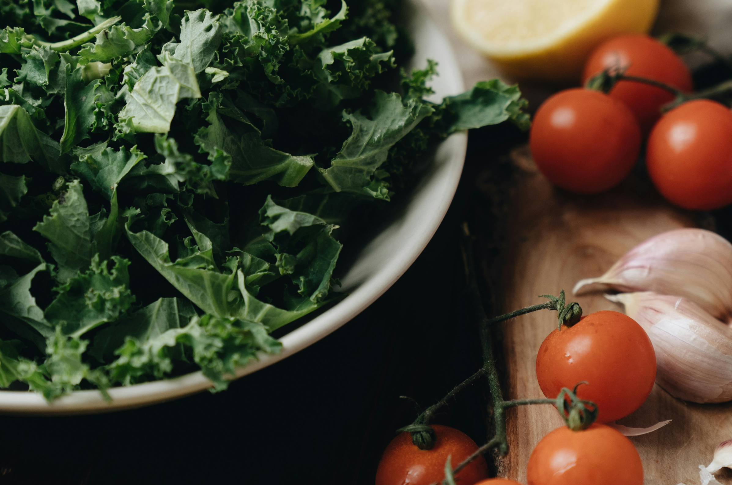 Pro Chefly winter prep ingredients with fresh kale in a bowl beside cherry tomatoes and garlic cloves on a wooden board, ready for slicing with Japanese Damascus knives.