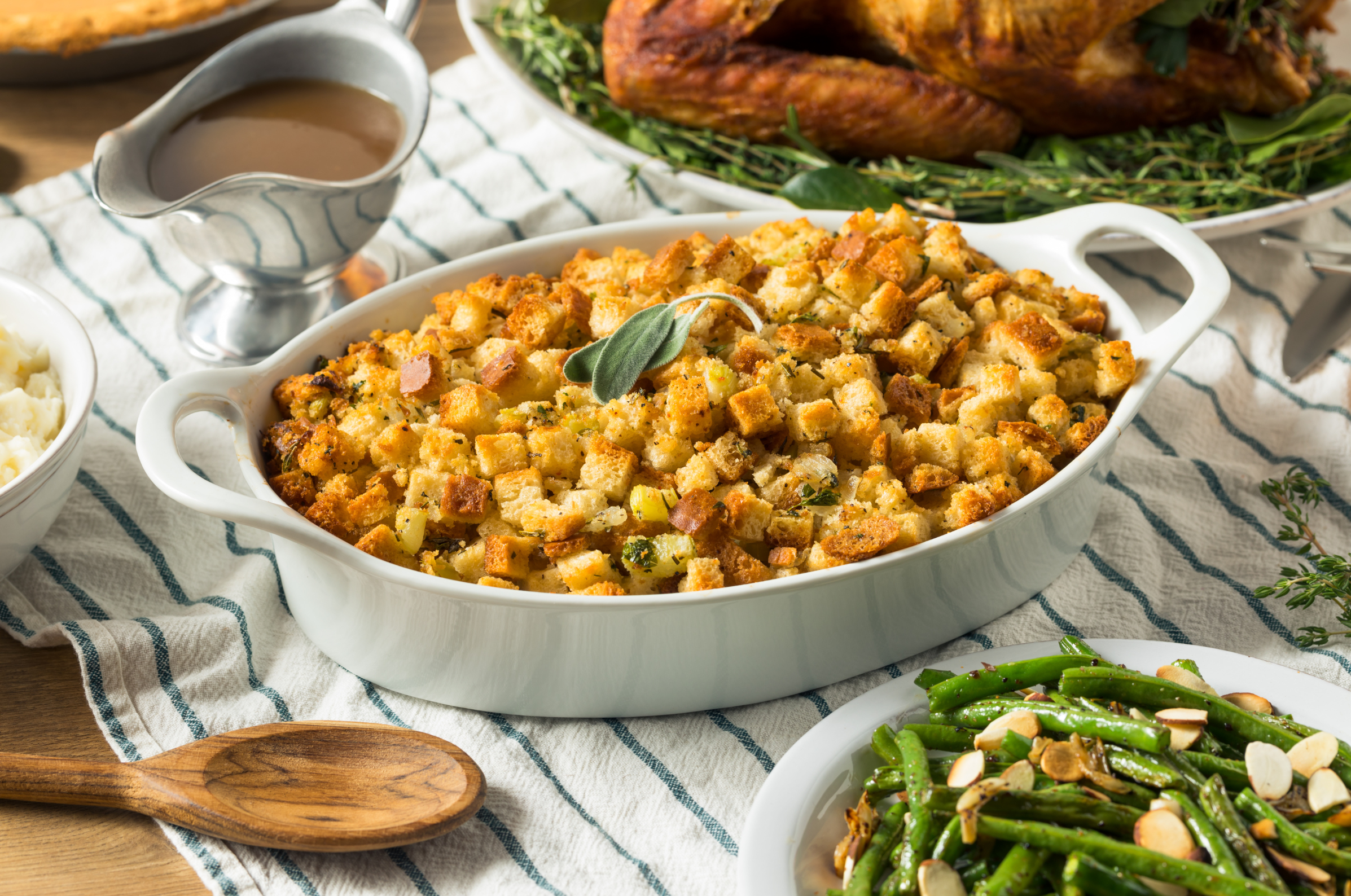 Golden baked stuffing in a white casserole dish topped with fresh sage, surrounded by classic holiday sides on a striped kitchen towel.