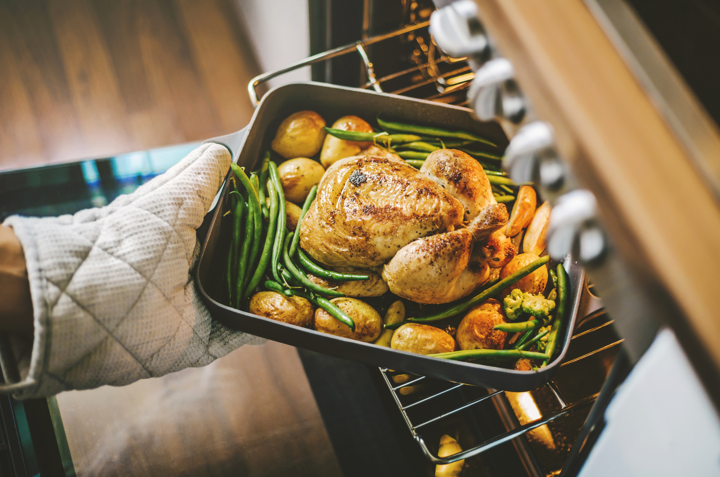 Image of **Alt text:** Roast chicken with potatoes and green beans being pulled from the oven with an oven mitt.