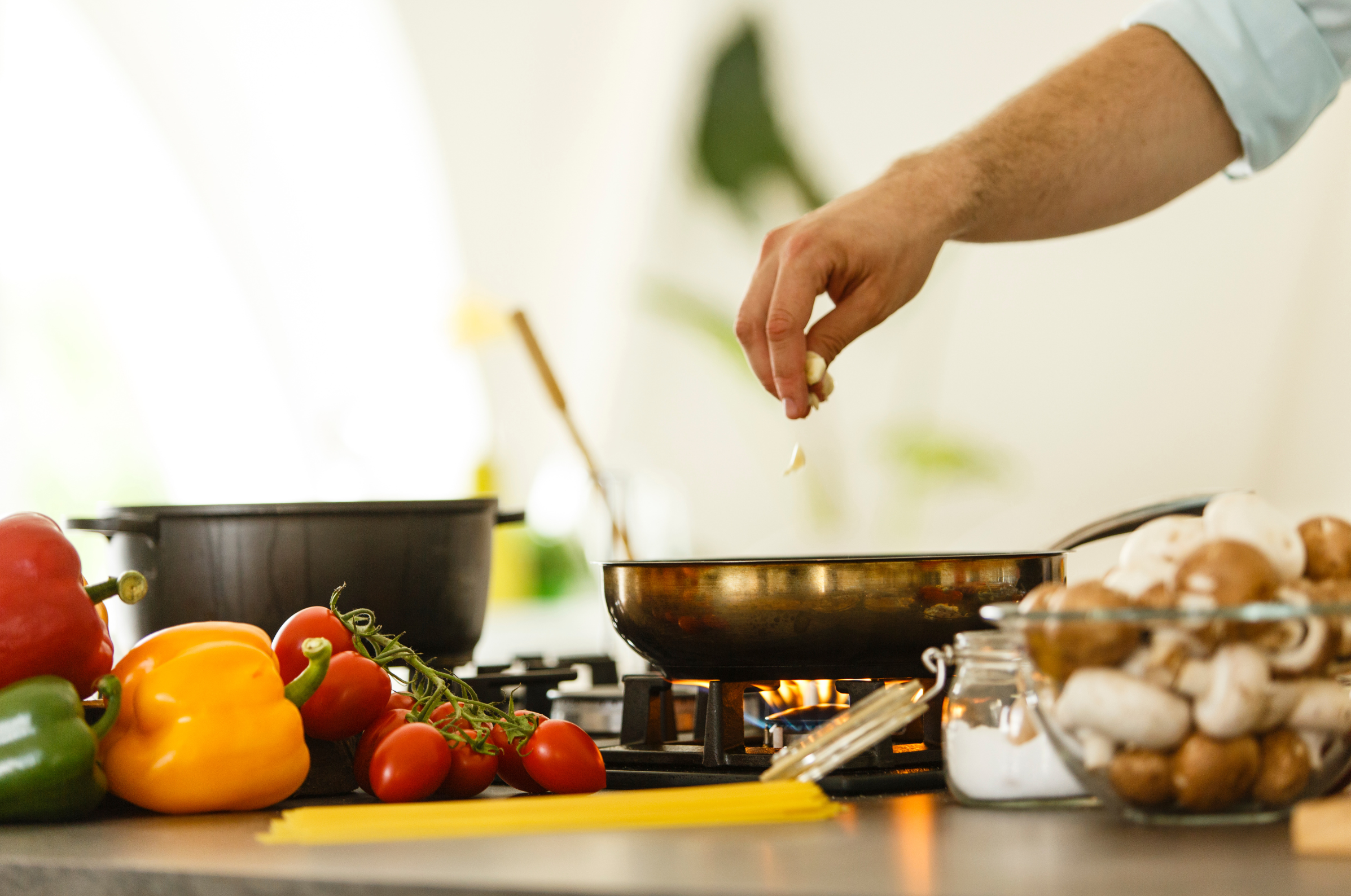 Image of **Alt text:** Hand adding garlic to a pan on the stove with peppers, tomatoes, mushrooms, and pasta on the counter.