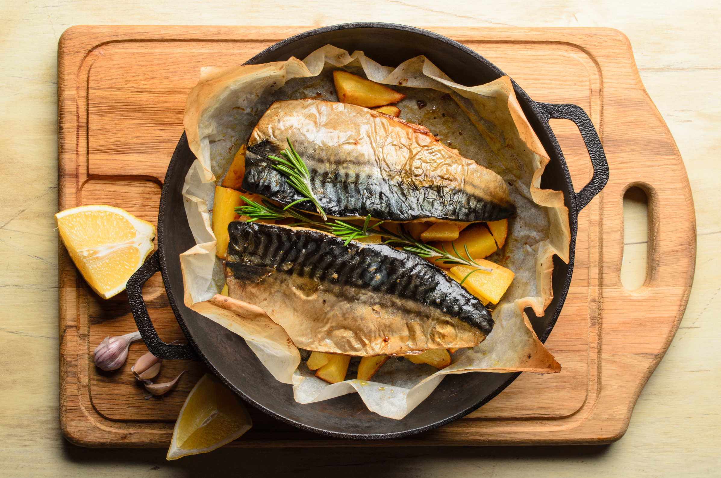 Baked mackerel fillets with rosemary and potatoes in a cast-iron pan with lemon and garlic on a wooden board.