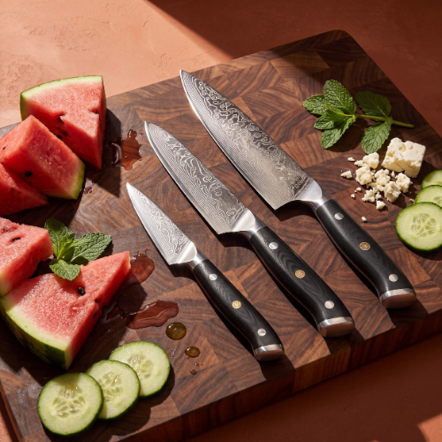 Image of Pro Chefly 3-piece Damascus knife set with black riveted handles, on a walnut board with watermelon, cucumber, mint and feta