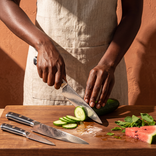 Image of Pro Chefly 3-piece Damascus knife set with black riveted handles, slicing cucumber on a walnut board with watermelon and mint