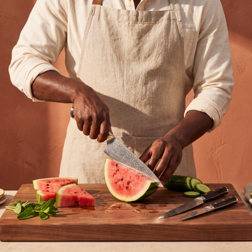 Image of Pro Chefly 3-piece Damascus knife set with black riveted handles, slicing watermelon on a walnut board with cucumber and mint